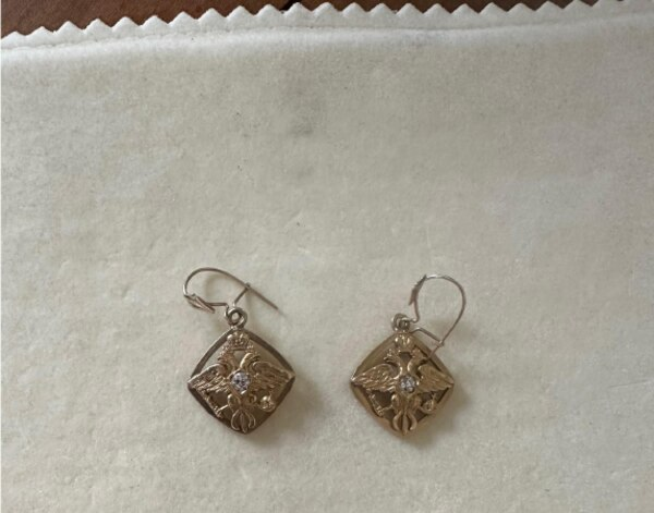 A pair of gold earrings made from family cufflinks, each with a crest motif and a small white stone