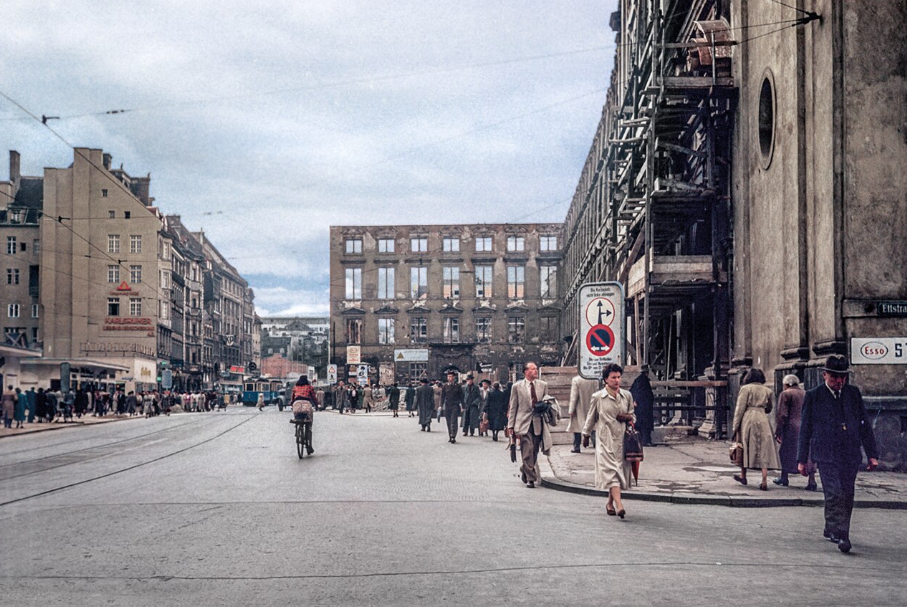 A postwar Munich street with scaffolding on a damaged building and pedestrians in period dress